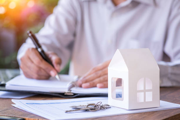 Personne qui signe un document avec une petite maquette de maison en papier et des clefs au premier plan